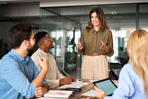 person stood up giving a talk to multiple people sat round a table 